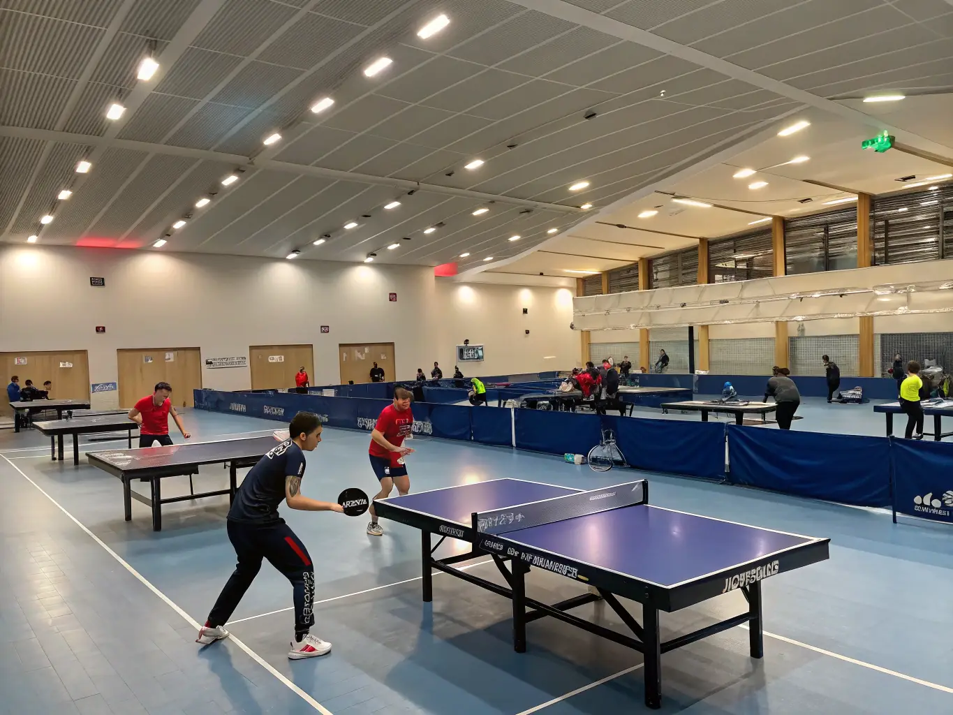 A vibrant indoor sports hall with table tennis tables set up, players in HCTT uniforms warming up for a local tournament.